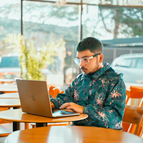 A young man focuses on his laptop in a bright Baguio cafe, wearing eyeglasses and a floral jacket.